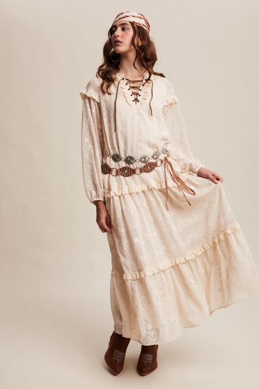 Model in ivory boho maxi dress with lace-up neckline, long puff sleeves, and tiered ruffle skirt, styled with concho belt, headscarf, and western boots.