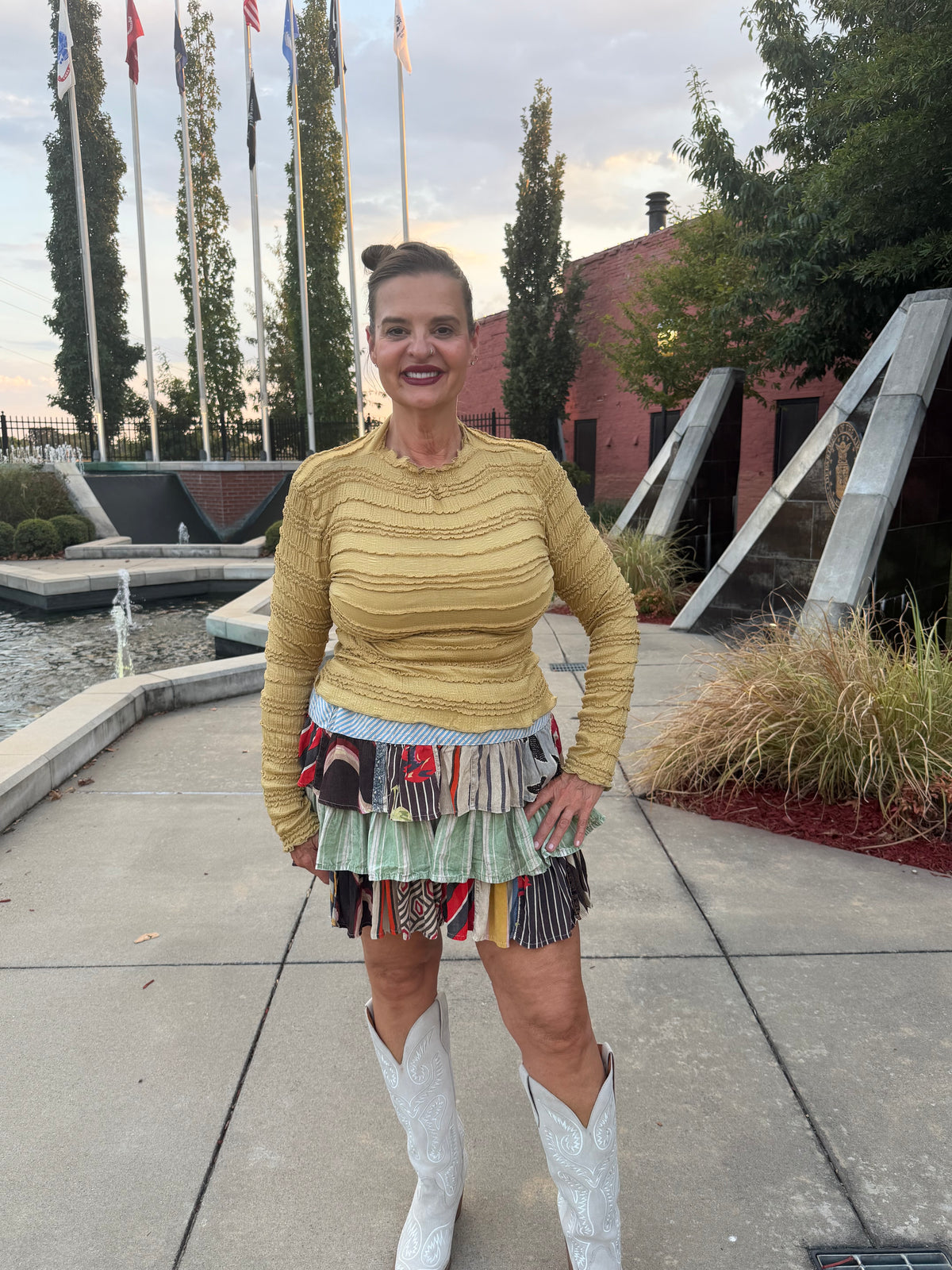 A woman stands outdoors on a paved walkway wearing a fitted mustard yellow ribbed long-sleeve top, a highly textured multicolor patchwork fringe skirt, and white cowboy boots.