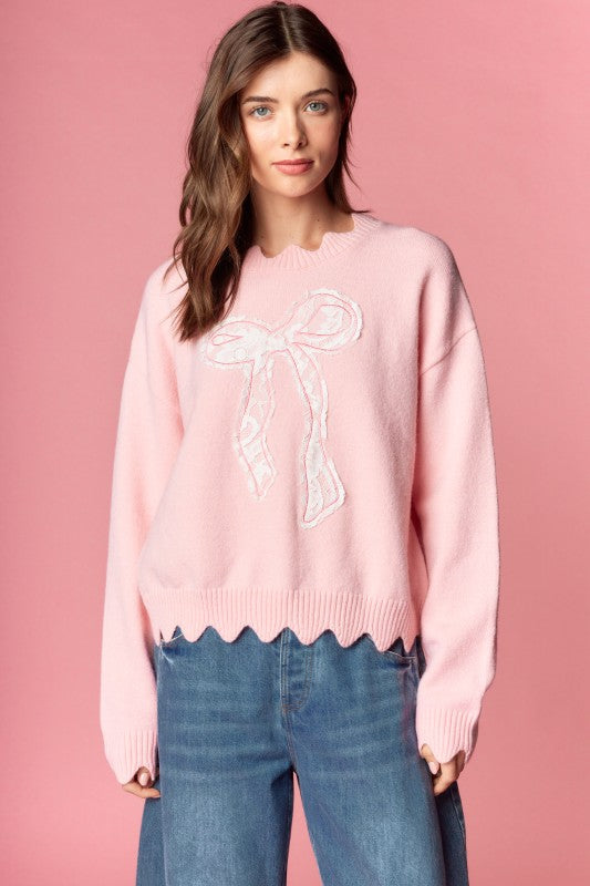 Pink knit sweater with white lace bow applique, relaxed fit, ribbed cuffs and scalloped hem, three-quarter front view on model.