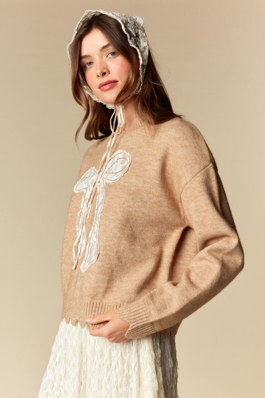 Camel knit sweater with white lace bow applique, relaxed fit, ribbed cuffs and scalloped hem, three-quarter front view on model.