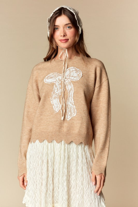 Camel knit sweater with white lace bow applique, relaxed fit, ribbed cuffs and scalloped hem, three-quarter front view on model.