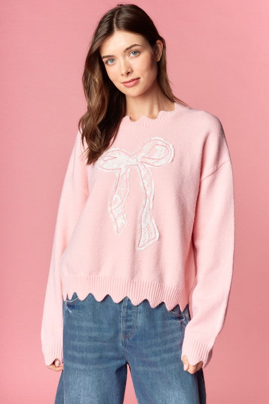 Pink knit sweater with white lace bow applique, relaxed fit, ribbed cuffs and scalloped hem, three-quarter front view on model.
