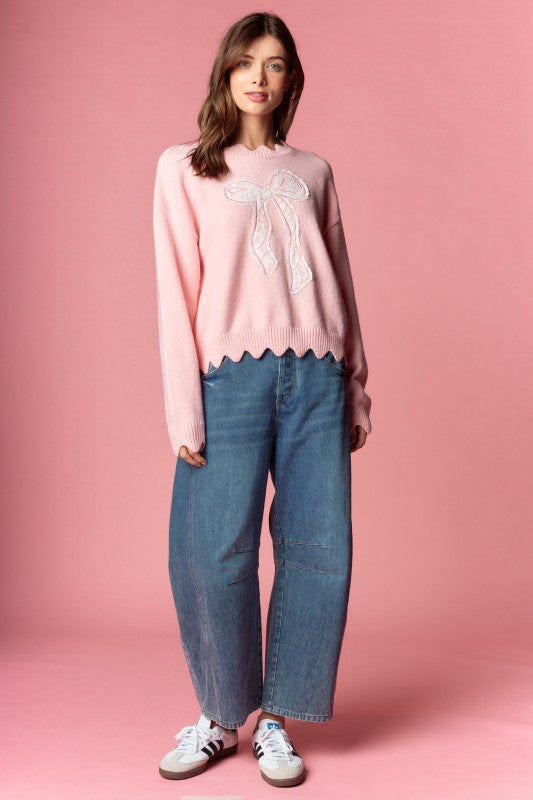 Pink knit sweater with white lace bow applique, relaxed fit, ribbed cuffs and scalloped hem, three-quarter front view on model.