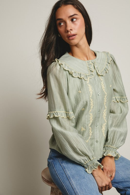 Model wearing sage green embroidered blouse with ruffled Peter Pan collar, button front, puff long sleeves and smocked cuffs, paired with tan trousers.
