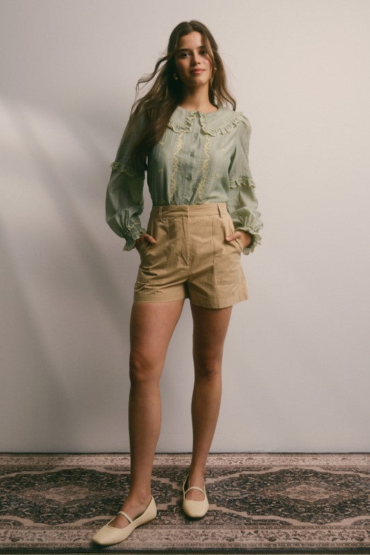Model wearing sage green embroidered blouse with ruffled Peter Pan collar, button front, puff long sleeves and smocked cuffs, paired with tan trousers.