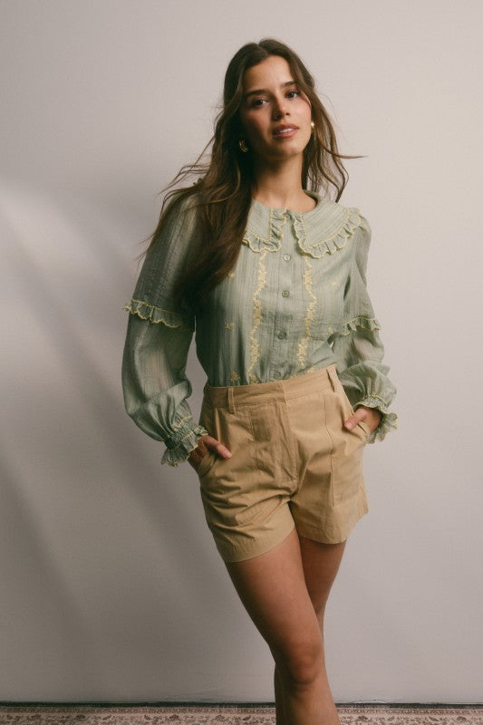 Model wearing sage green embroidered blouse with ruffled Peter Pan collar, button front, puff long sleeves and smocked cuffs, paired with tan trousers.