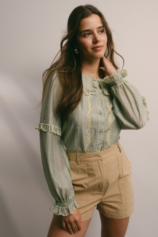 Model wearing sage green embroidered blouse with ruffled Peter Pan collar, button front, puff long sleeves and smocked cuffs, paired with tan trousers.