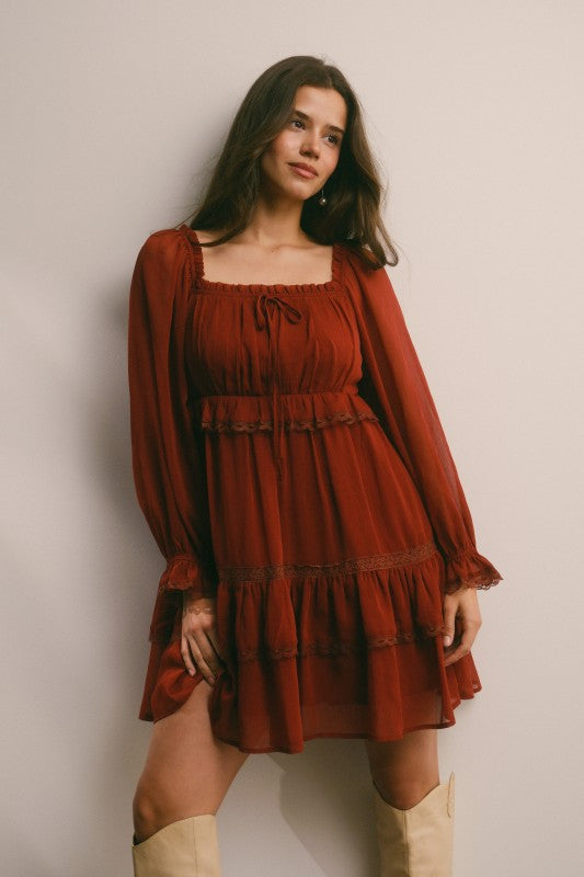 Model wearing rust chiffon babydoll mini dress with square neckline, tie-front, long sheer sleeves, tiered ruffle hem, and side pockets.