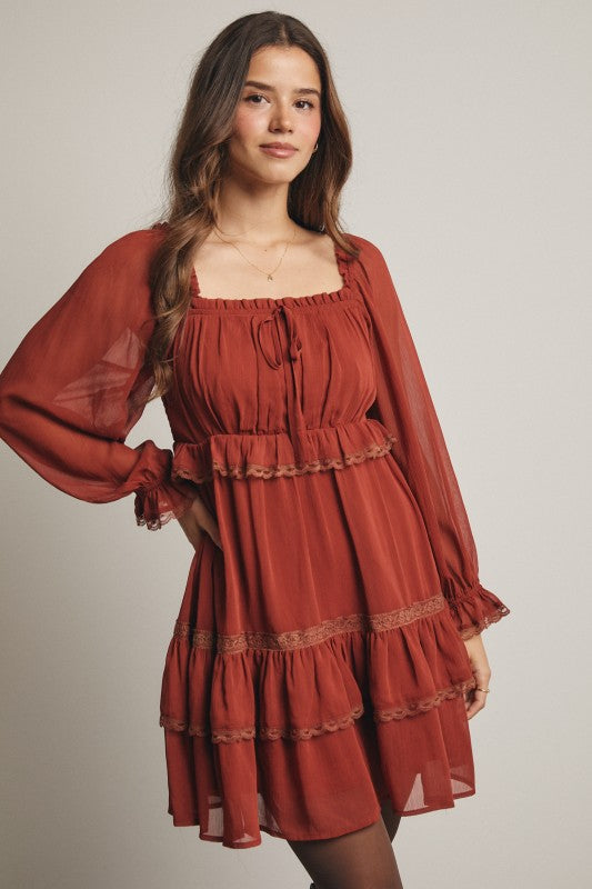 Model wearing rust chiffon babydoll mini dress with square neckline, tie-front, long sheer sleeves, tiered ruffle hem, and side pockets.