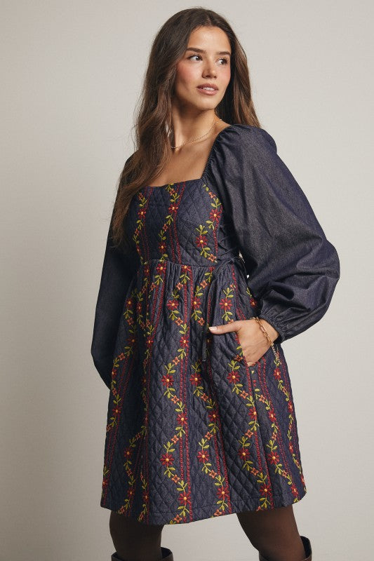 Model in a navy quilted dress with colorful floral embroidery and corset-laced waist, denim puff sleeves; side view, knee-length.