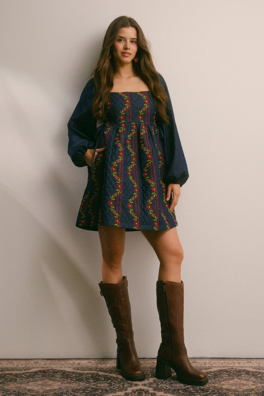 Model in a navy quilted dress with colorful floral embroidery and corset-laced waist, denim puff sleeves; side view, knee-length.