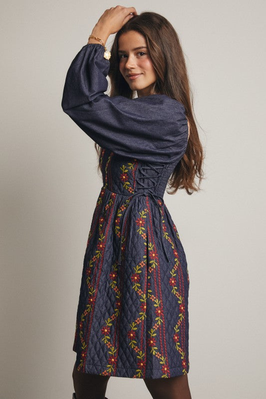 Model in a navy quilted dress with colorful floral embroidery and corset-laced waist, denim puff sleeves; side view, knee-length.