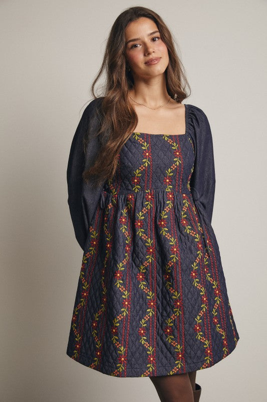 Model in a navy quilted dress with colorful floral embroidery and corset-laced waist, denim puff sleeves; side view, knee-length.