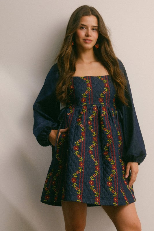 Model in a navy quilted dress with colorful floral embroidery and corset-laced waist, denim puff sleeves; side view, knee-length.