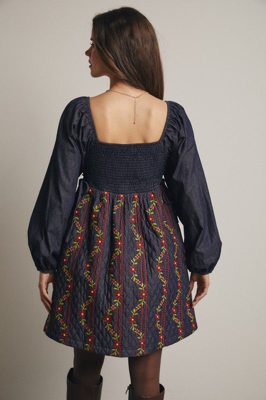 Back view of a navy quilted mini dress with colorful floral embroidery, smocked back bodice, and denim puff sleeves; model wearing boots.