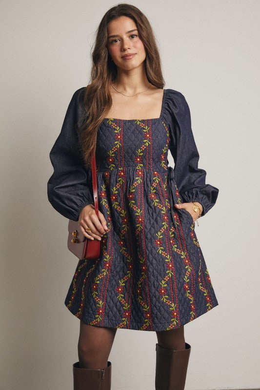 Model in a navy quilted dress with colorful floral embroidery and corset-laced waist, denim puff sleeves; side view, knee-length.