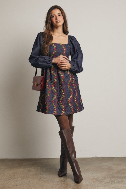 Model in a navy quilted dress with colorful floral embroidery and corset-laced waist, denim puff sleeves; side view, knee-length.