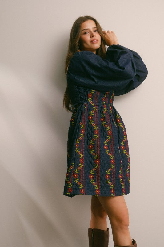 Model in a navy quilted dress with colorful floral embroidery and corset-laced waist, denim puff sleeves; side view, knee-length.
