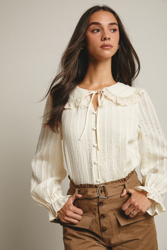 Ivory tie-neck blouse with ruffled Peter Pan collar, covered buttons, textured stripe fabric, and long puff sleeves.