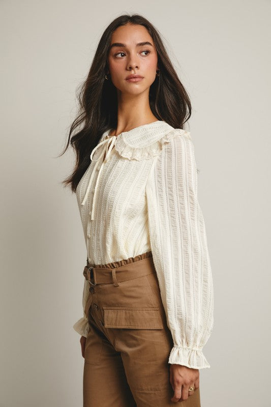 Ivory tie-neck blouse with ruffled Peter Pan collar, covered buttons, textured stripe fabric, and long puff sleeves.