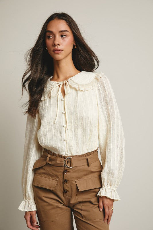 Ivory tie-neck blouse with ruffled Peter Pan collar, covered buttons, textured stripe fabric, and long puff sleeves.