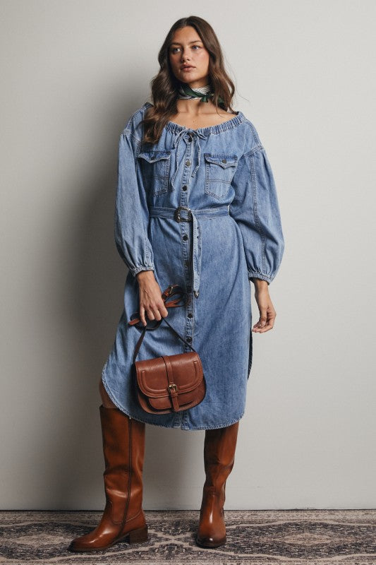 Woman wearing a denim dress with brown boots and a bag against a plain background