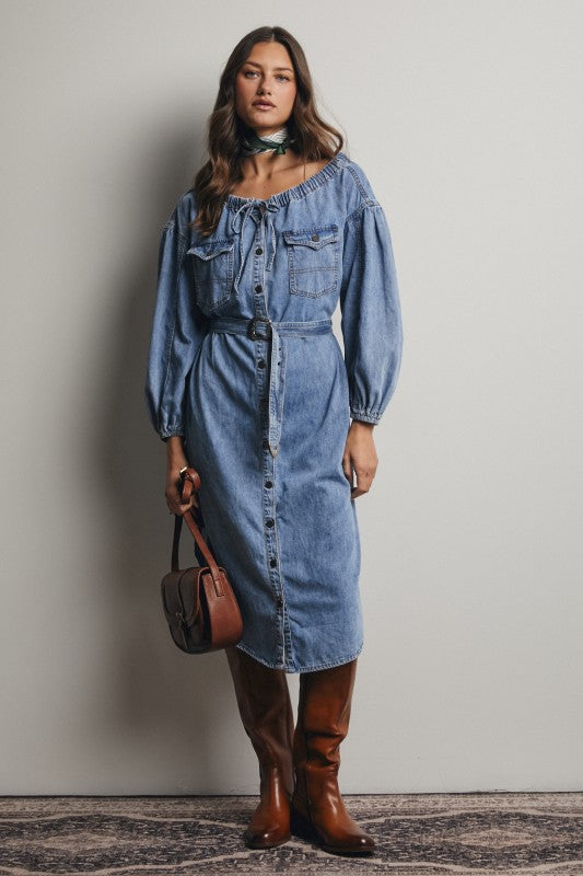 Woman wearing a denim dress with long sleeves and brown boots, holding a brown handbag against a plain background.