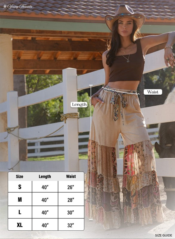 The Dreamer Tiered Wide Leg Pants styled with casual top - whimsical eclectic boho pants with a size chart in front of a white fence.