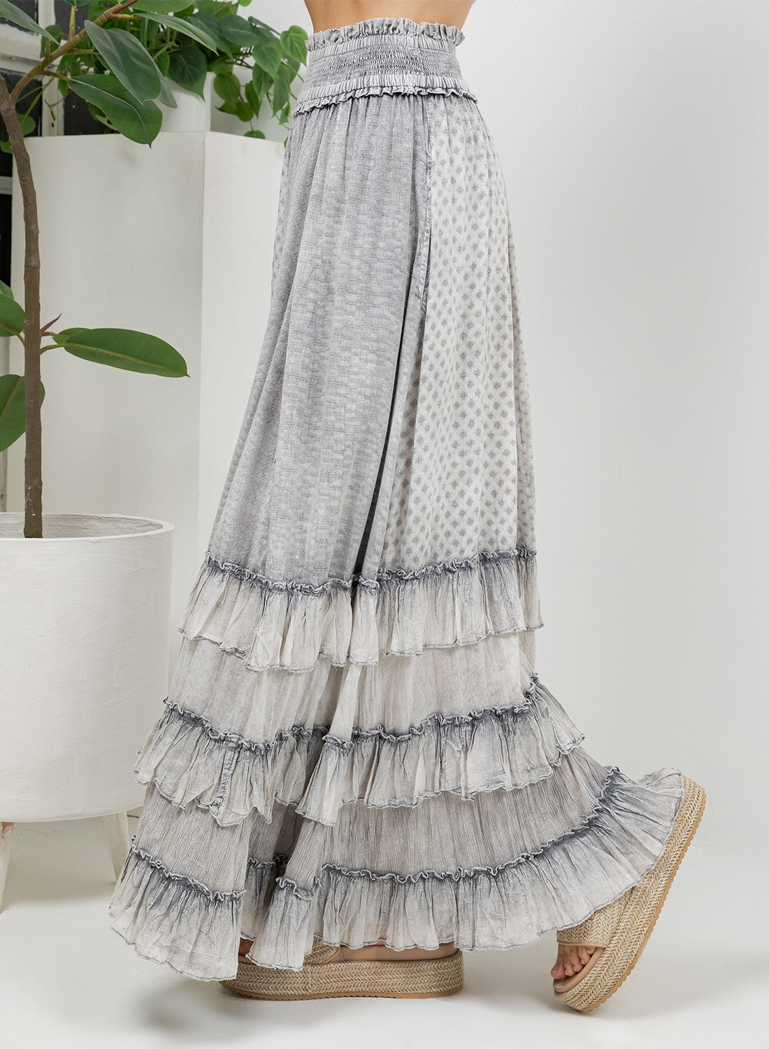 A long tiered maxi skirt in a light gray and white patterned fabric, shown on a model from the waist down wearing espadrille sandals in a bright studio setting.