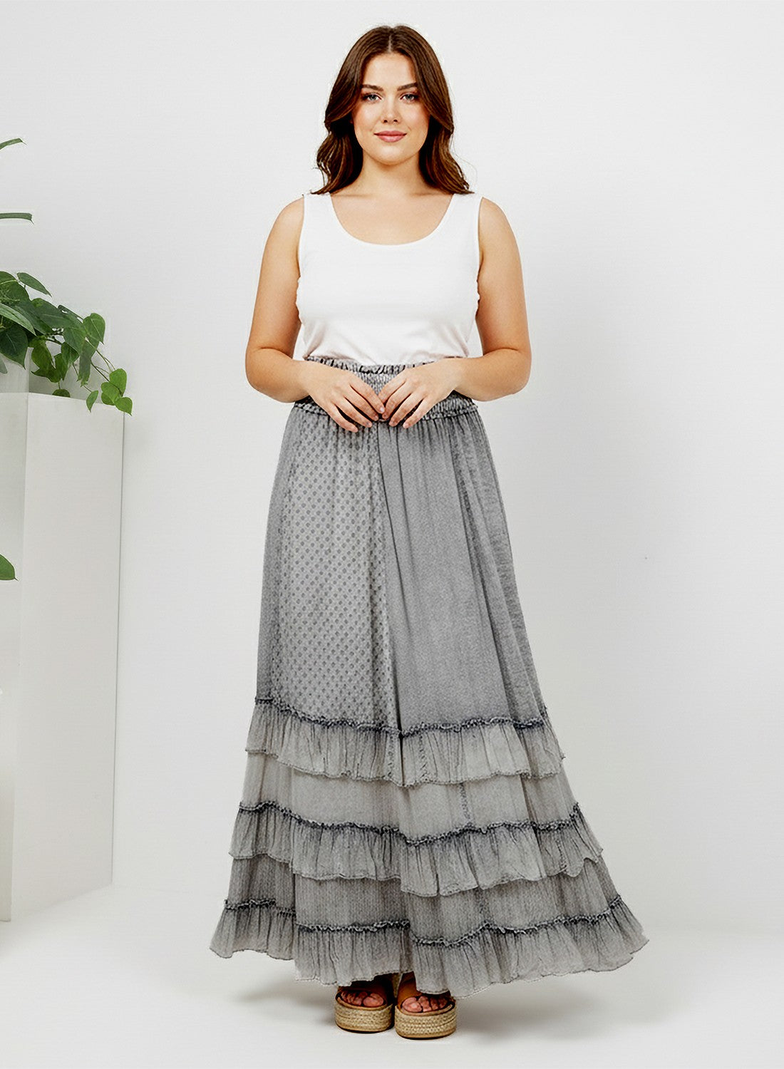 A woman modeling a long tiered gray skirt with a small polka dot pattern, paired with a white sleeveless top and brown espadrille sandals, standing against a white studio background.