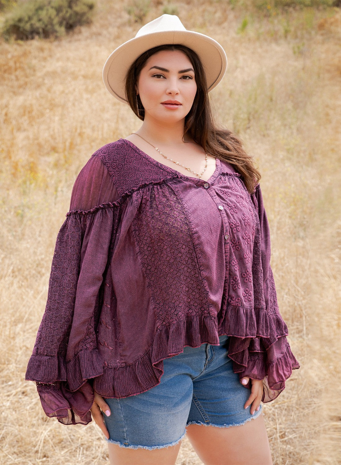 A woman wearing a purple bohemian-style lace top with ruffled sleeves and a white wide-brim hat, standing in a dry grassy field.