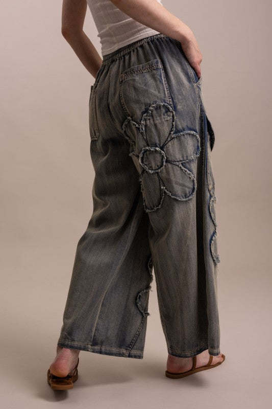Jeans with large floral applique details on the back, shown from the waist down against a plain beige studio background