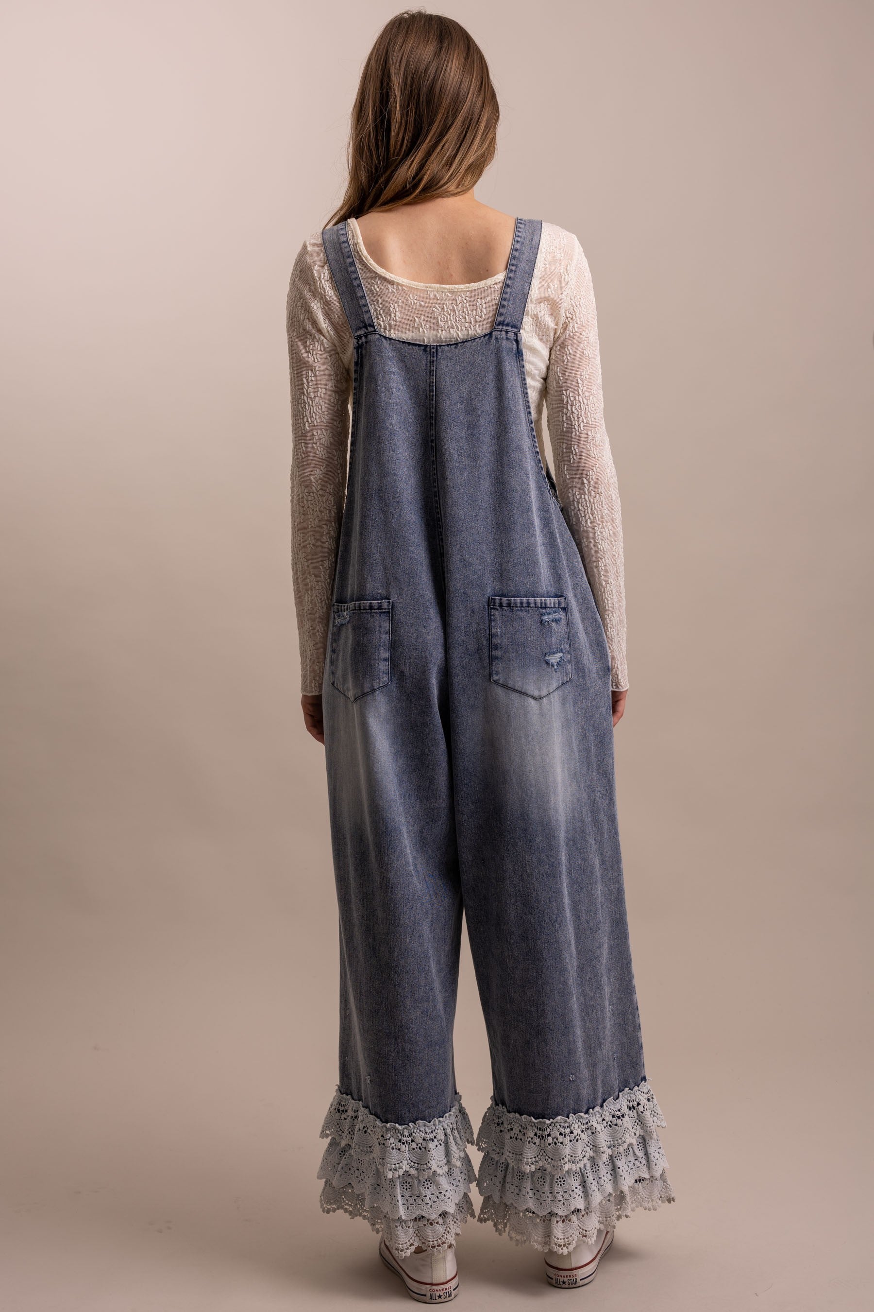 Back view of distressed wide leg denim overall with tiered lace ruffle hem and adjustable straps, two back patch pockets