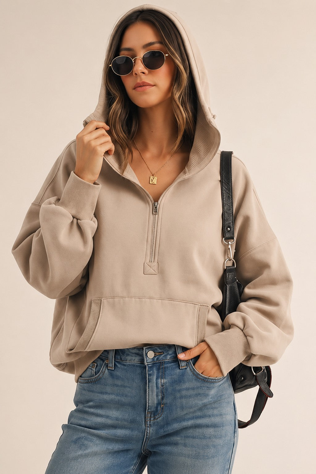 A woman wearing a beige half-zip hoodie with a front kangaroo pocket, paired with blue jeans, sunglasses, and a black shoulder bag, set against a plain beige background.