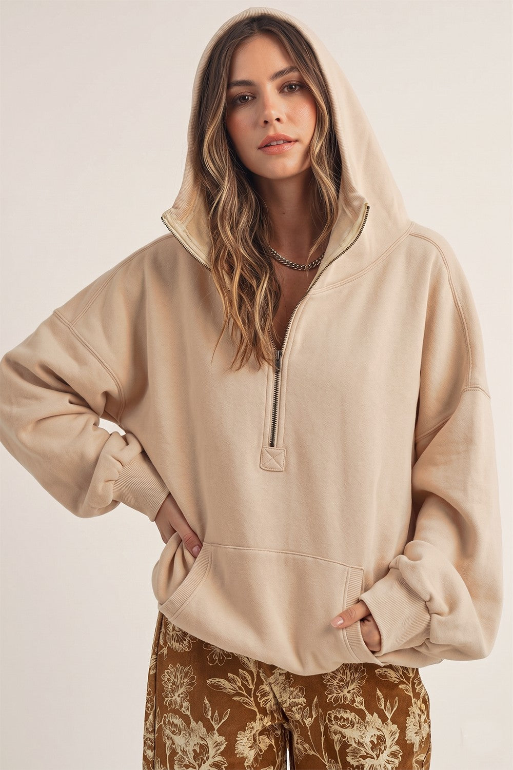 A woman wearing an oversized beige half-zip hoodie with a drawstring hood and front kangaroo pockets, paired with brown floral pants, against a plain light background.