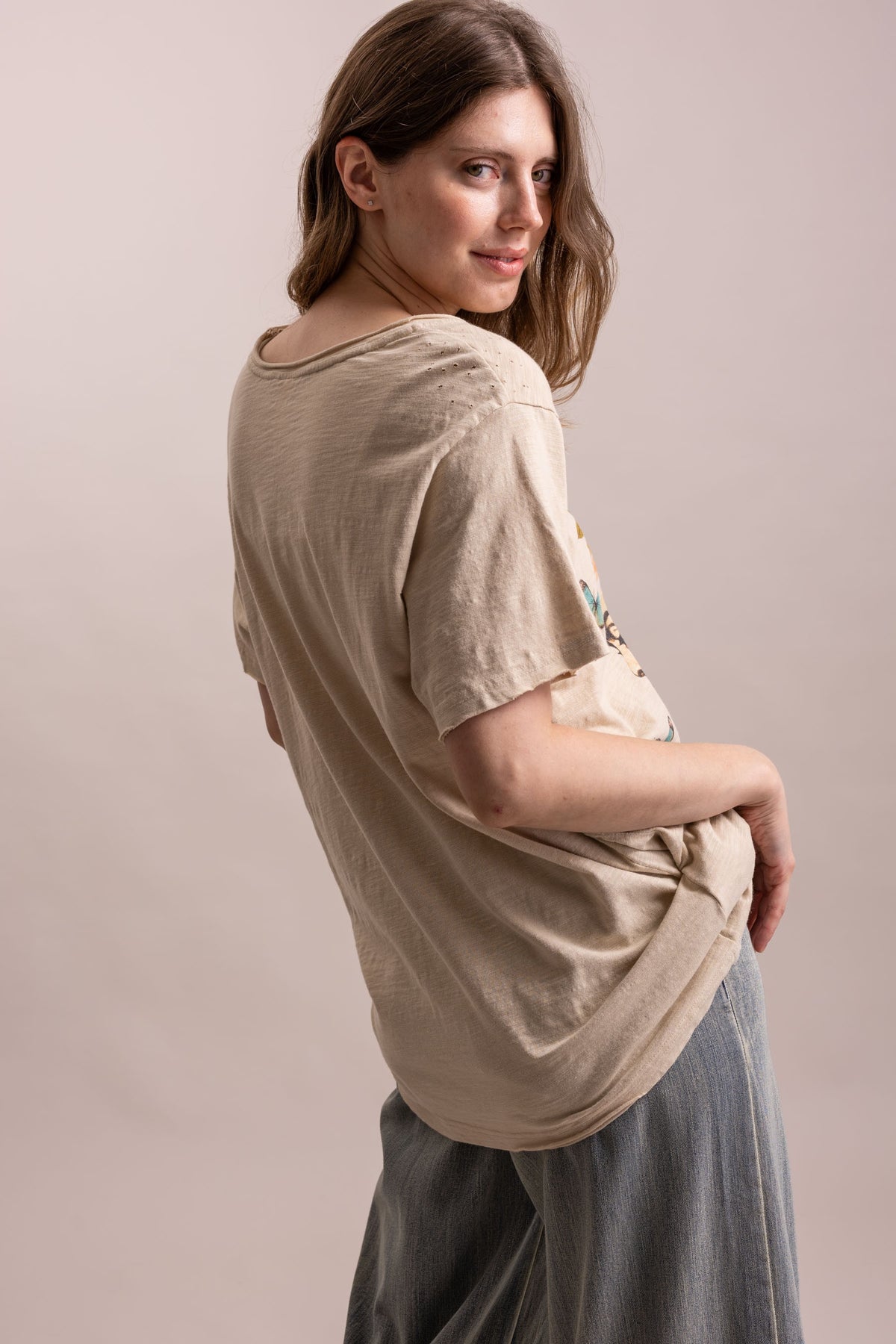 A woman wearing a beige short-sleeve t-shirt and gray wide-leg pants, photographed from a side-back angle against a solid light beige studio background.