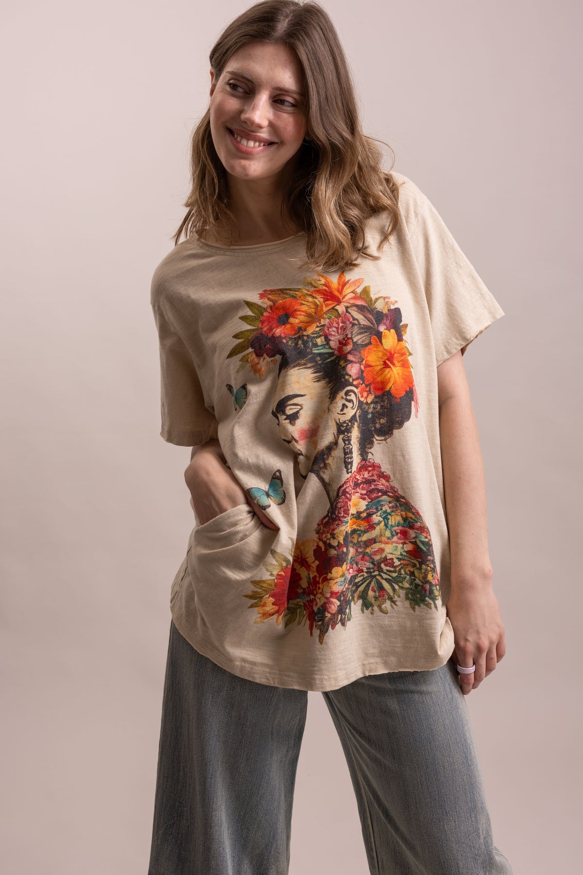 A woman wearing a beige oversized graphic t-shirt with a colorful floral and portrait print, paired with gray wide-leg jeans, against a solid light beige background.