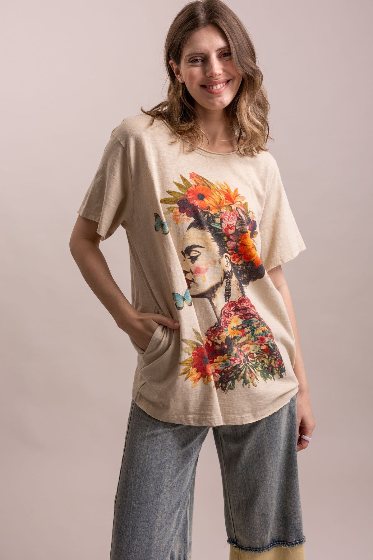 A woman modeling a beige oversized graphic t-shirt featuring a colorful floral portrait of Frida Kahlo, paired with wide-leg denim jeans, against a solid light beige studio background.
