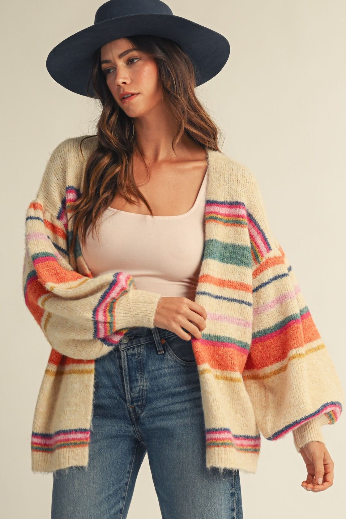 A woman wearing a cream-colored fuzzy knit cardigan with colorful horizontal stripes in pink, orange, green, and blue, paired with a beige tank top, blue jeans, and a black wide-brim hat against a light beige background.