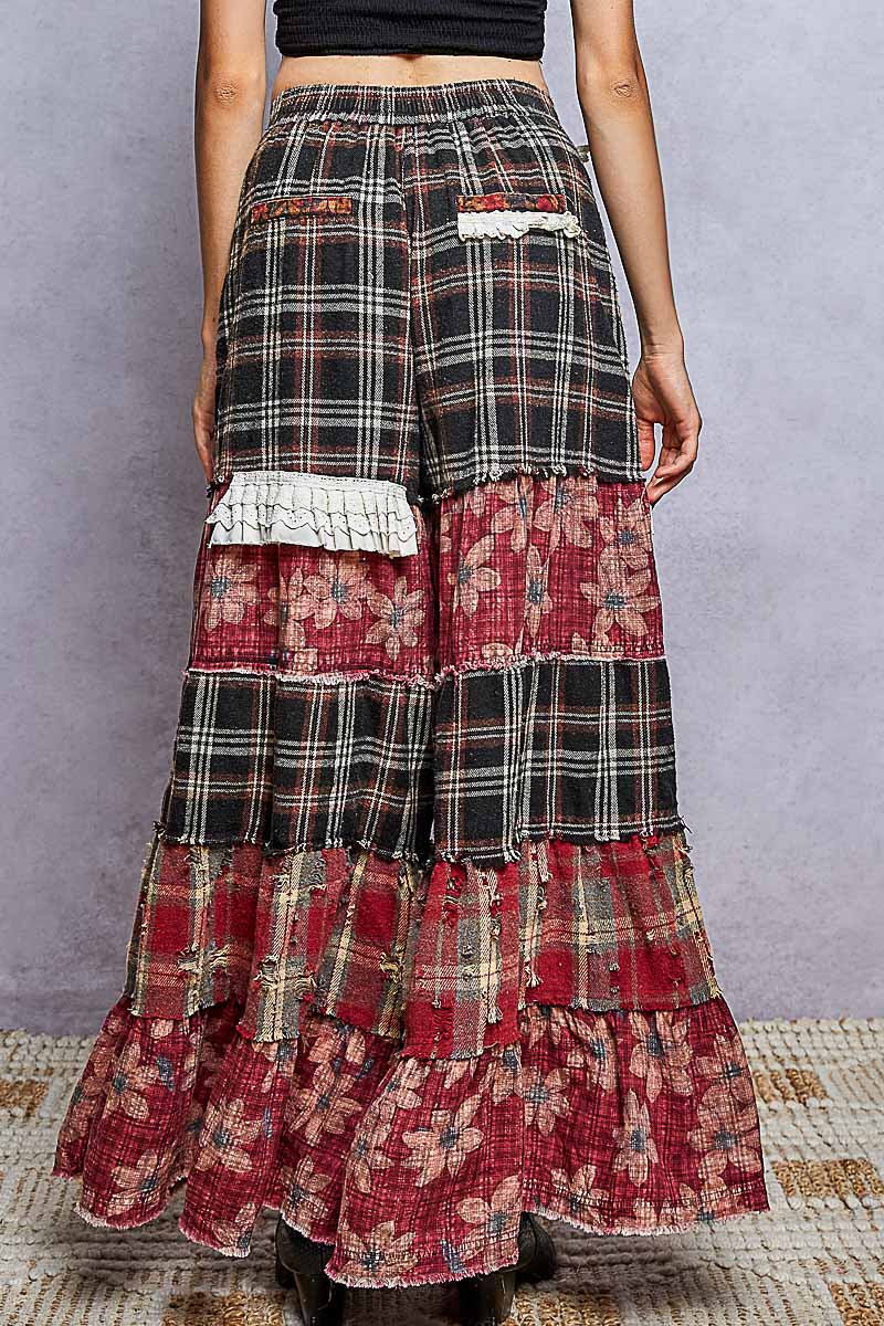 A person shown from the back wearing a long, tiered patchwork skirt with plaid and floral fabric panels, photographed against a textured gray wall and woven rug.