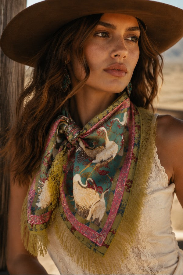 A woman wearing a brown wide-brim hat and a cream lace top, styled with a patterned fringed scarf featuring swans and a brown hat in the background.