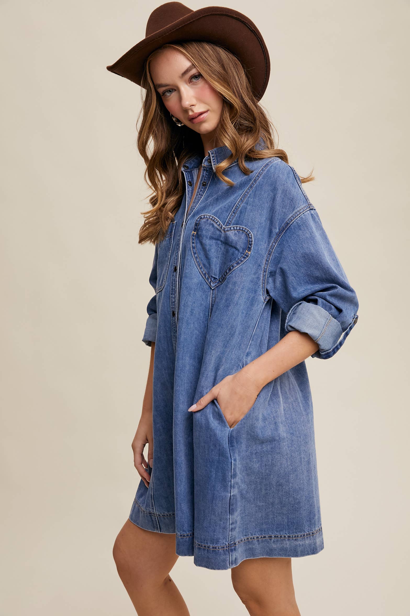 Side view- Medium-wash denim shirt dress with snap button front, heart-shaped chest pocket, rolled long sleeves, and a relaxed mini silhouette.