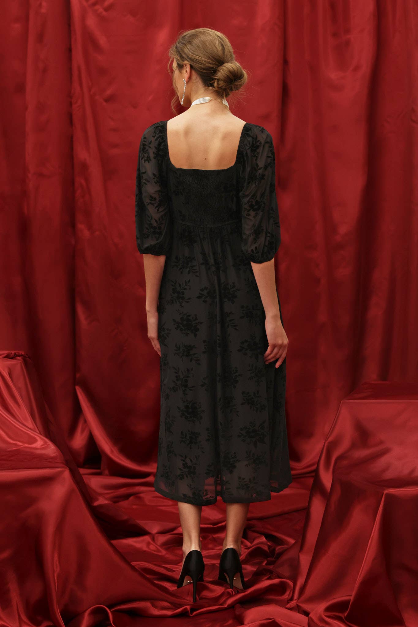 Rear view Model wearing a black velvet floral burnout midi dress with sweetheart neckline, twist-front bodice, and 3/4 puff sleeves with elastic cuffs, styled for an elegant holiday look against a red backdrop.