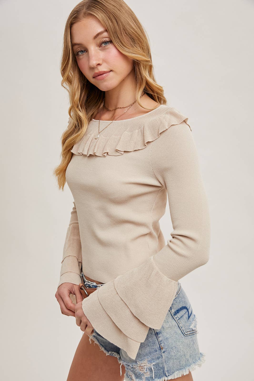 Beige ribbed sweater with ruffle neckline and tiered bell sleeves, paired with distressed denim shorts and a tan belt.