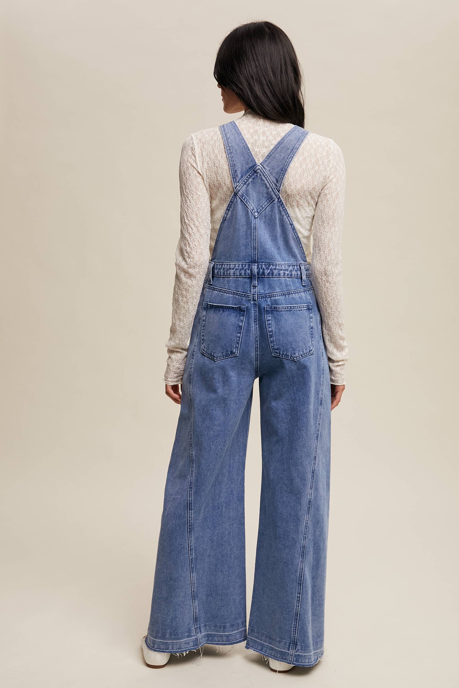 Back view- Light-wash wide-leg denim overalls with adjustable straps, front bib pocket, side buttons, and slant pockets, finished with a raw hem and layered over a cream long-sleeve top.