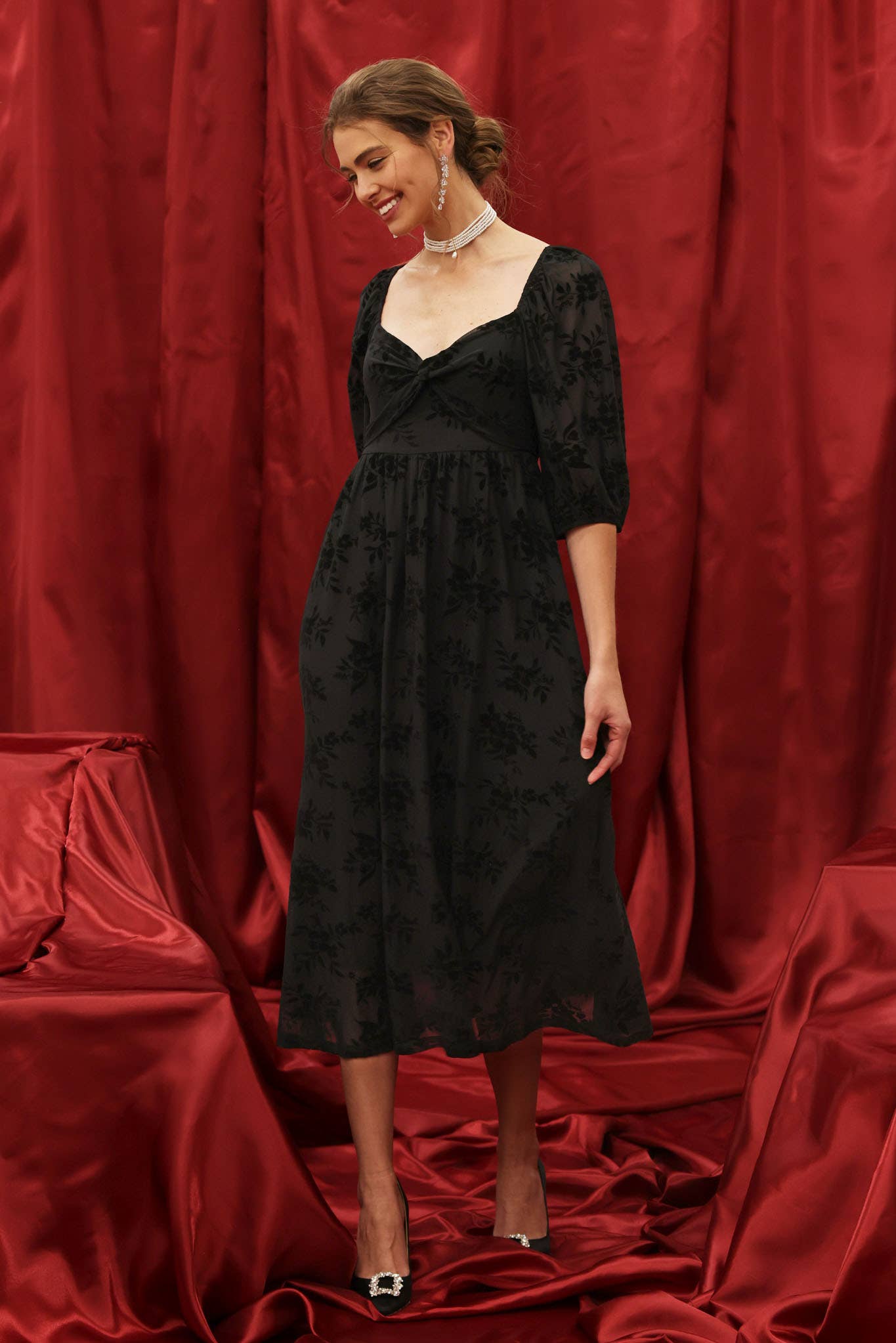 Model wearing a black velvet floral burnout midi dress with sweetheart neckline, twist-front bodice, and 3/4 puff sleeves with elastic cuffs, styled for an elegant holiday look against a red backdrop.