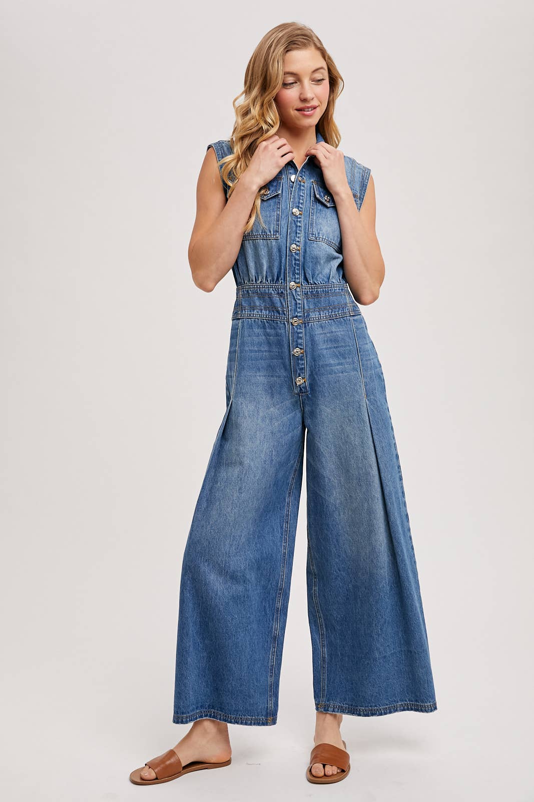 Sleeveless denim jumpsuit with collared button-front, chest flap pockets, and wide-leg palazzo silhouette, shown on model with slide sandals.