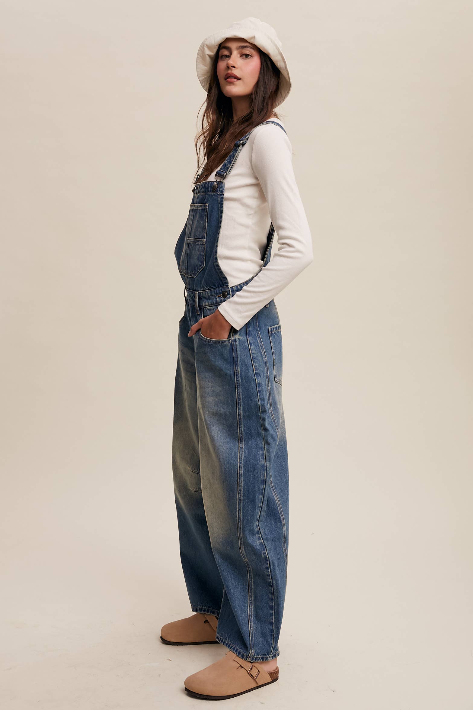 Side view- Vintage-inspired blue denim overalls with relaxed wide-leg fit, front chest pocket, and adjustable straps, styled with a white long-sleeve top and neutral clogs.