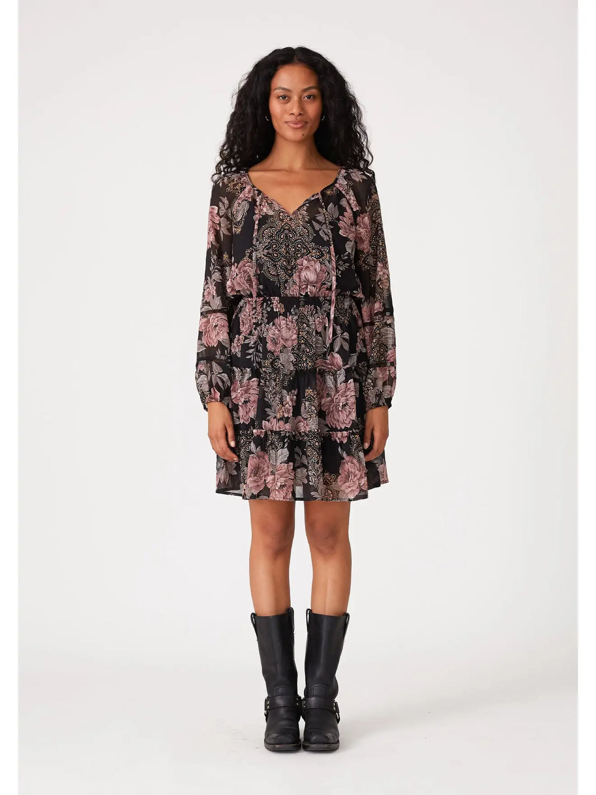 A woman modeling a long-sleeve tiered mini dress with a black base and pink floral pattern, paired with black knee-high boots, against a plain light gray studio background.