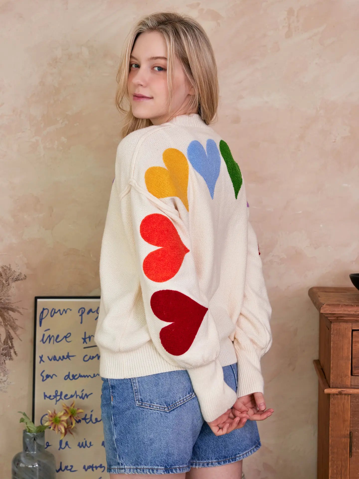 Person wearing a white cardigan with colorful heart patterns on the back, standing against a beige wall.